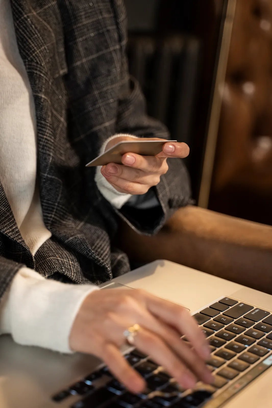 A woman in a checked blazer holding a bank card in one hand while typing on a laptop