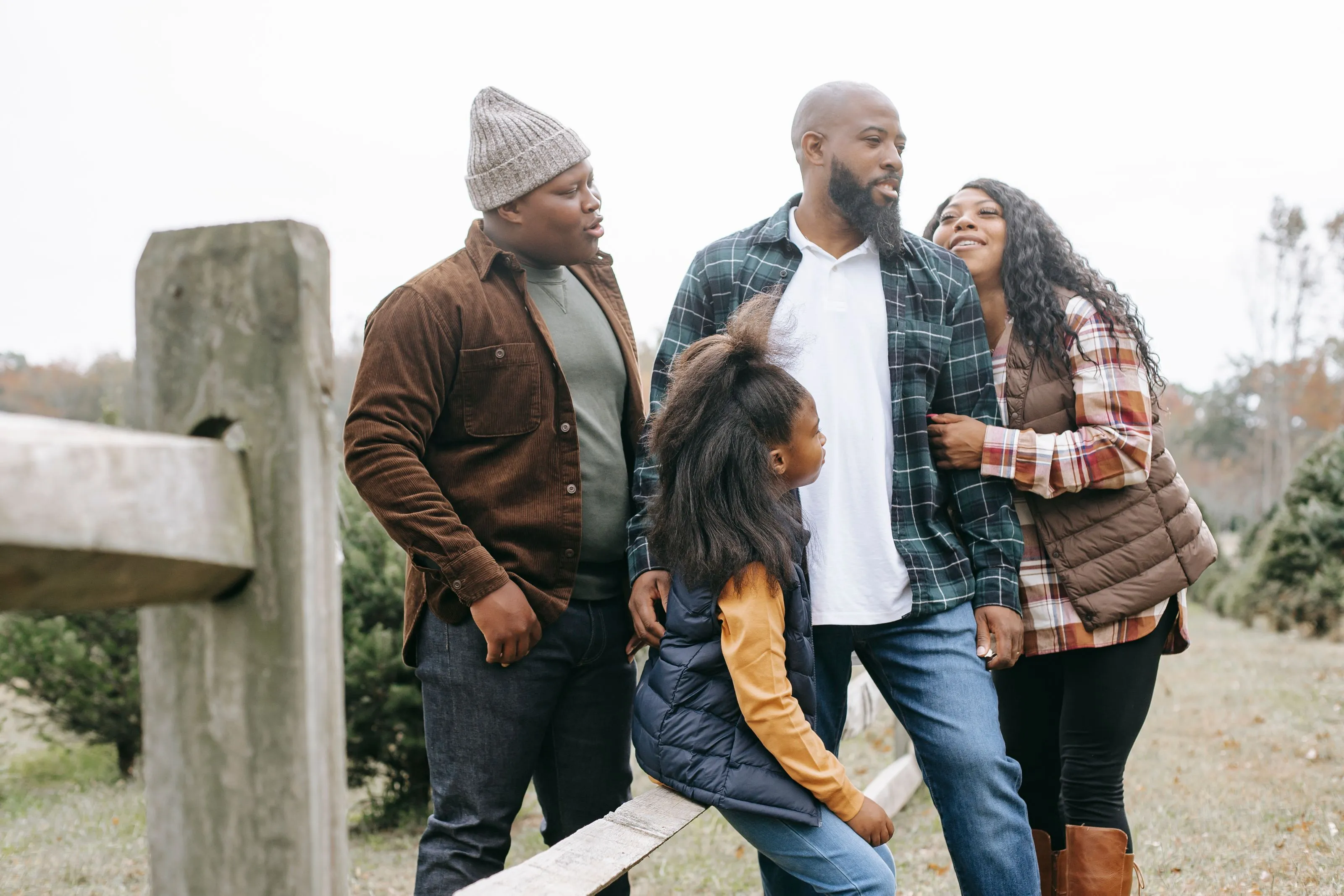 A Black family of four, three adults and a young girl, talking together beside a wooden fence outdoors