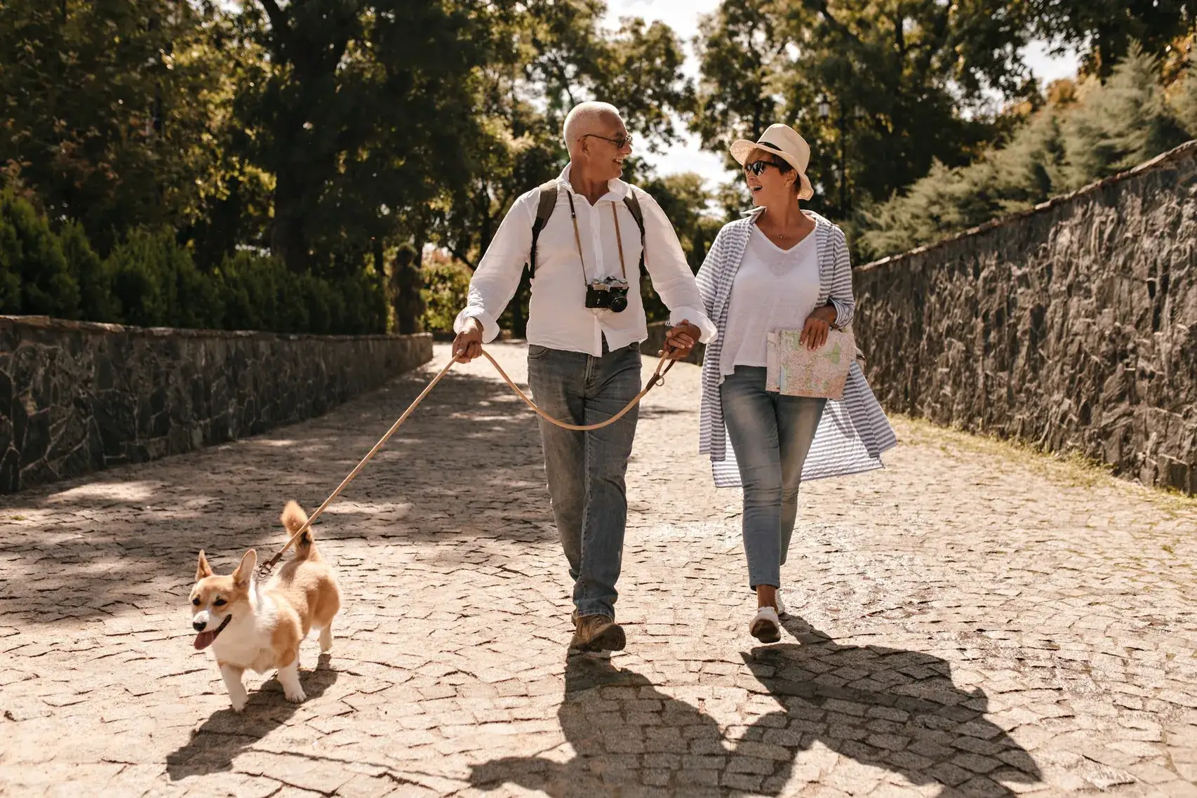 An older couple walking a corgi along a tree-lined path