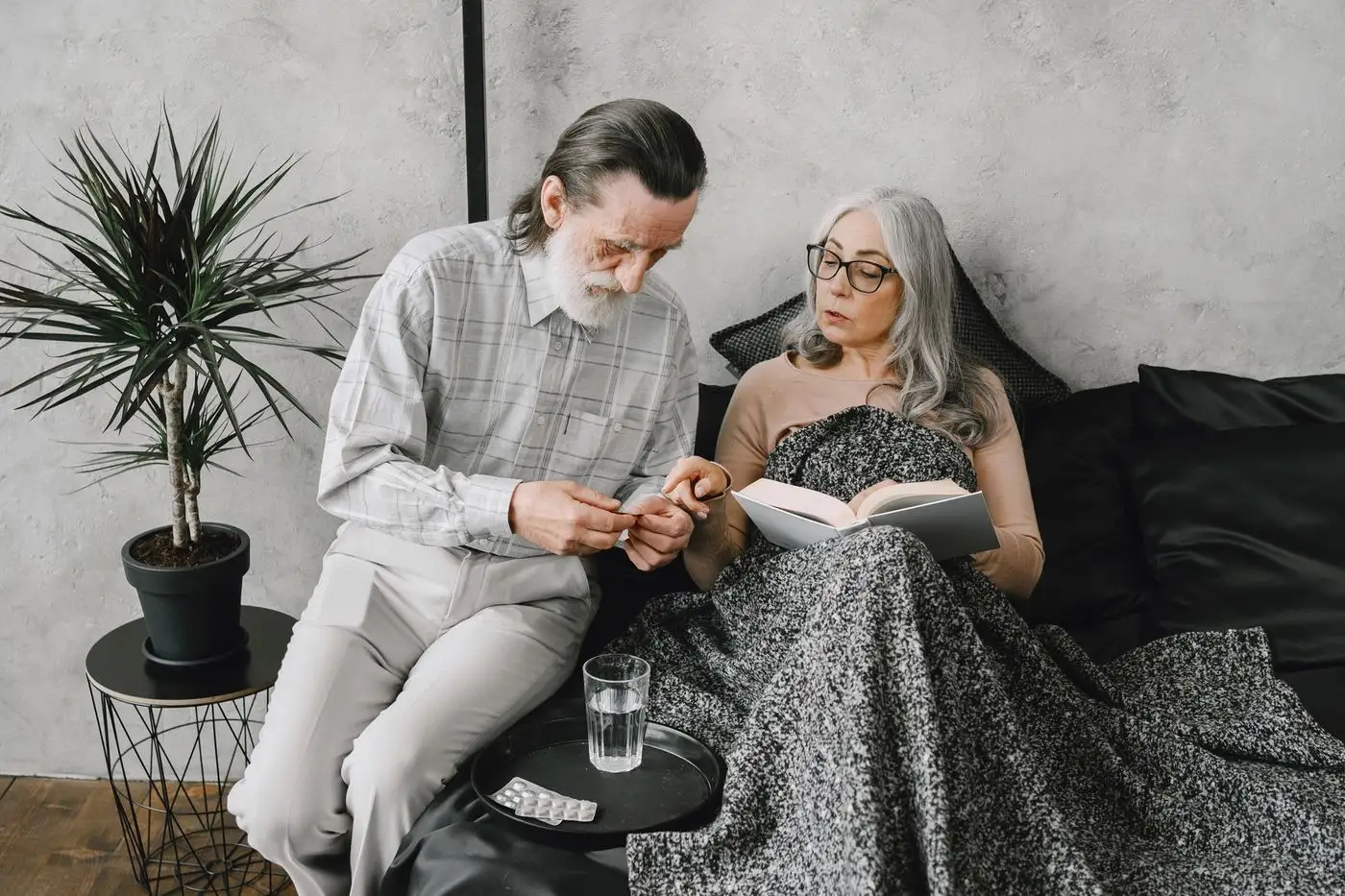 An older couple sitting together on a sofa, a glass of water and tablets on the table beside them
