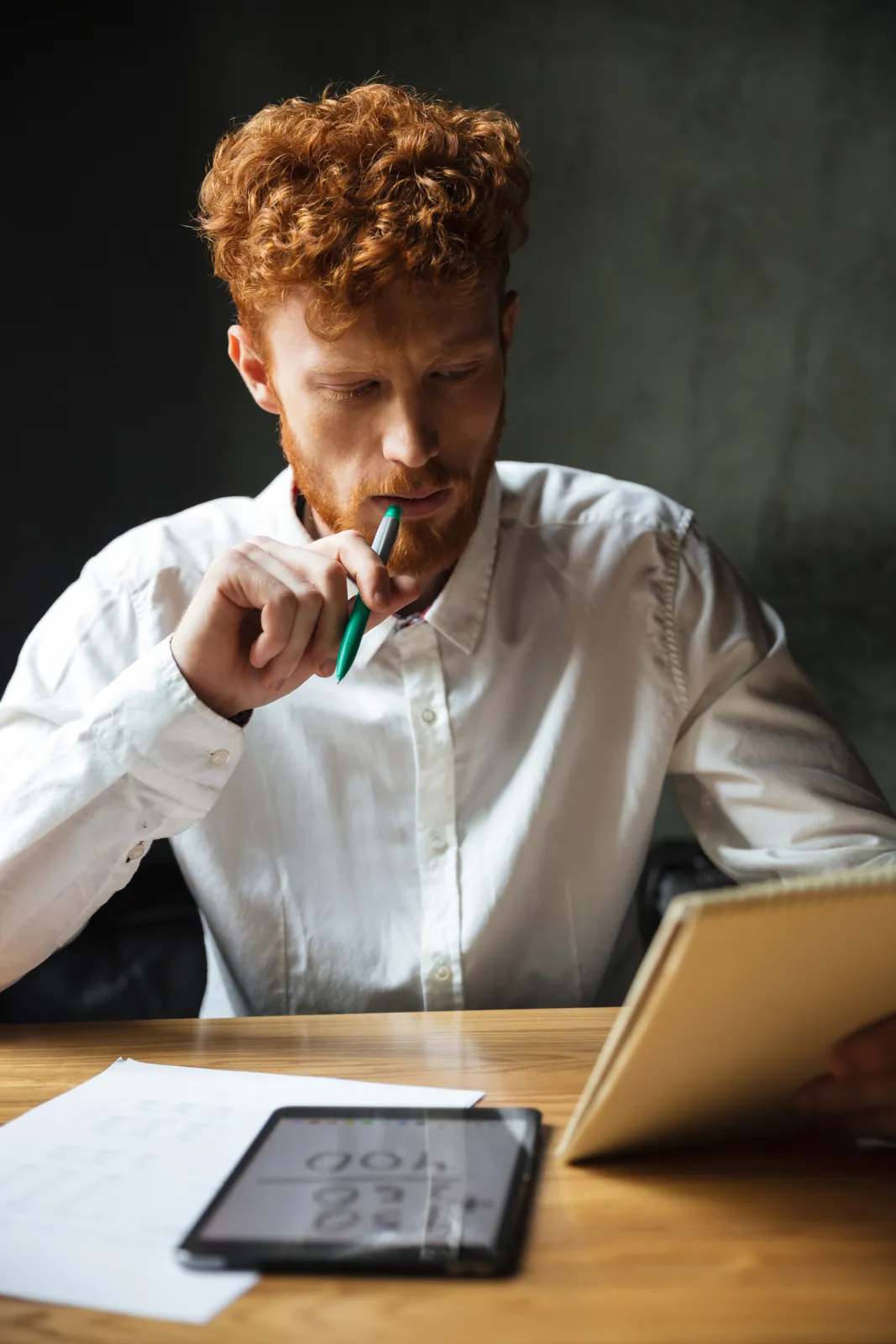 A young man at a desk pausing over an open notebook, a pen held to his lips