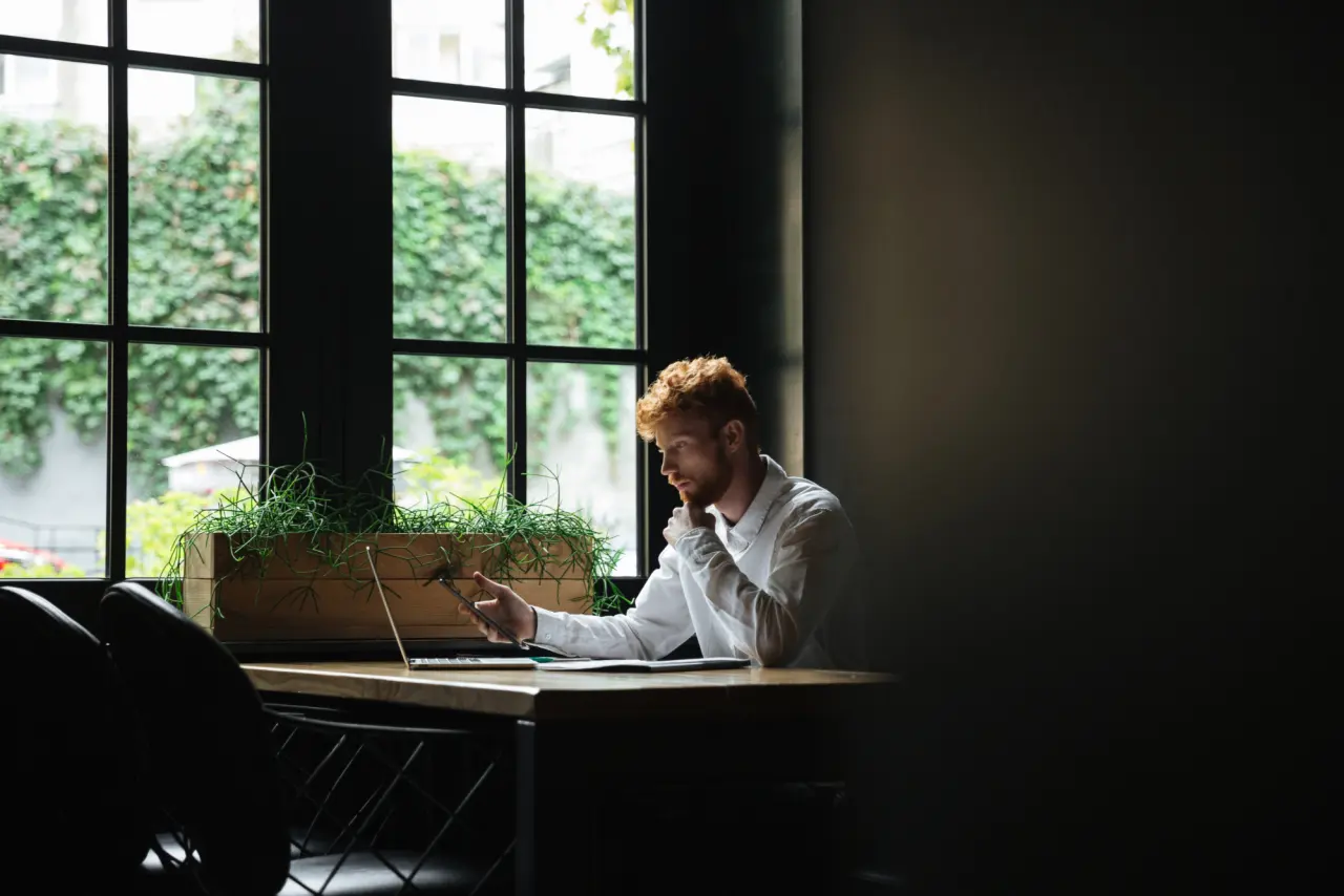 A man working at his desk beside a tall window, taking a phone call while reading from his laptop