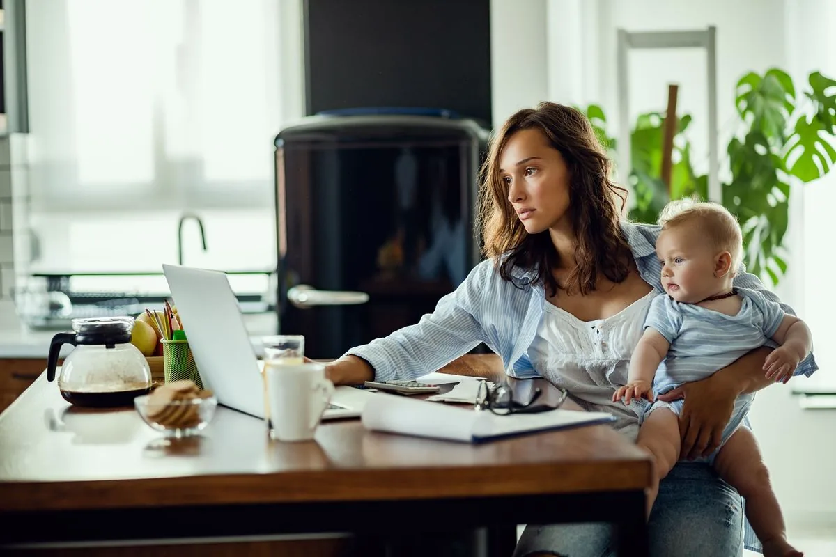 A woman working on a laptop at her kitchen table while holding her baby