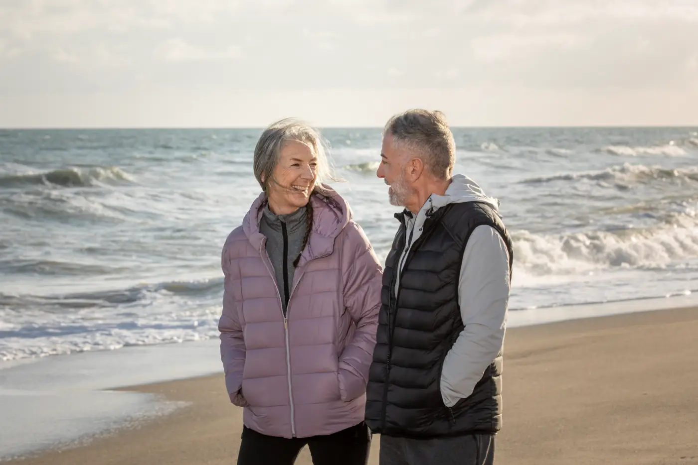 An older couple in winter coats smiling at each other on a windswept beach