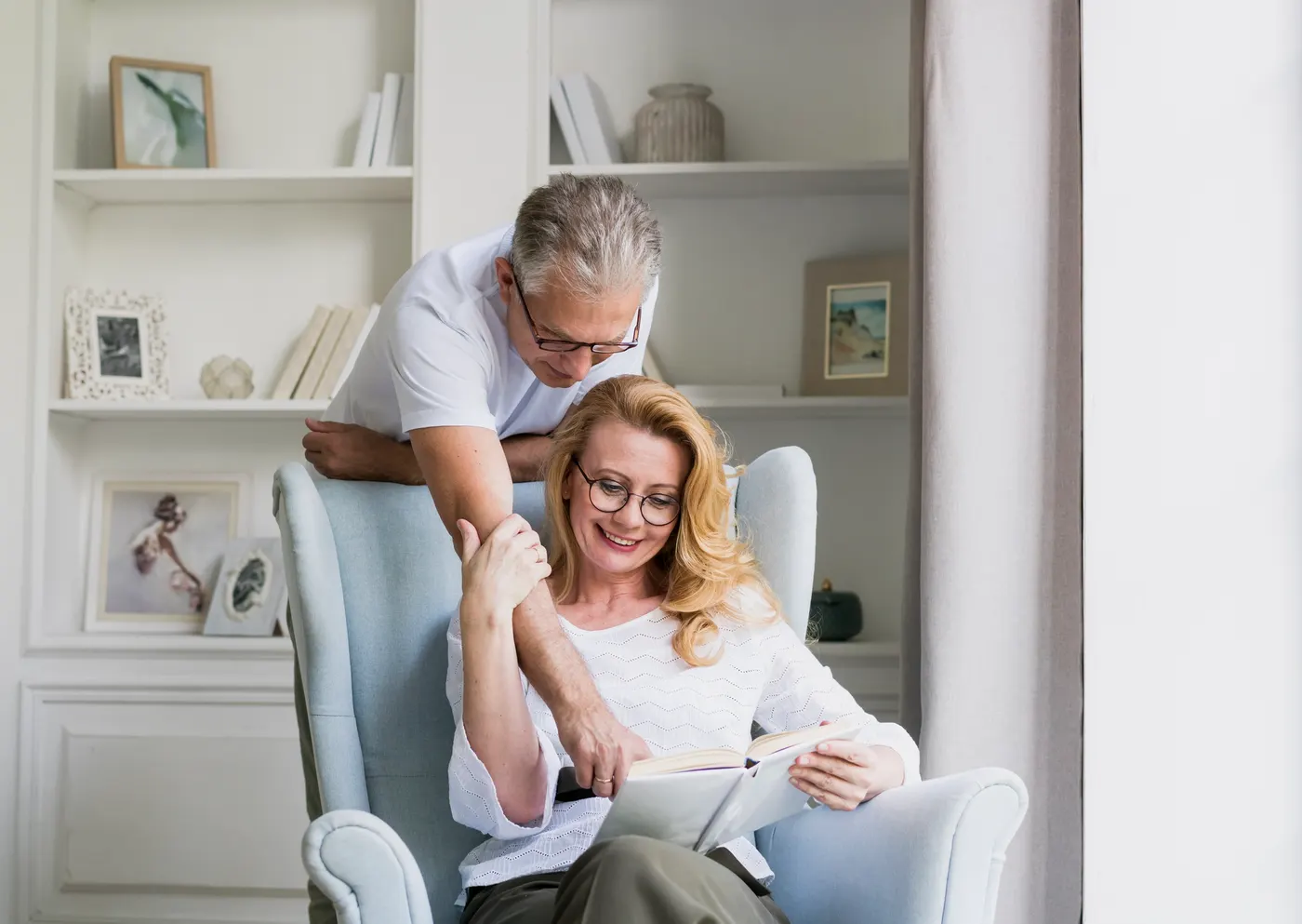 An older man leaning over an armchair to read a book with a woman