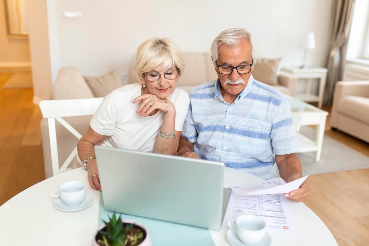 An older couple sitting together at their dining table, reading a printed statement and a laptop screen