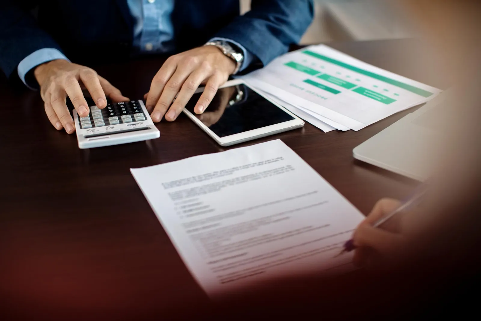 A person working through figures on a calculator beside a tablet and printed financial statements at a desk
