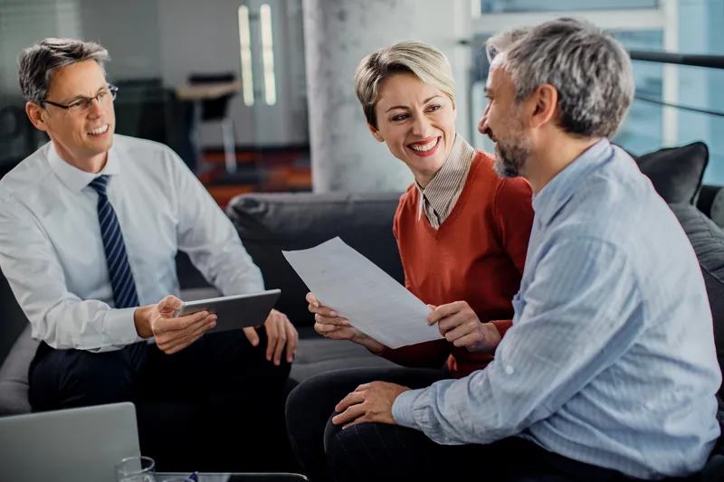 An adviser holding a tablet talking with a smiling couple on a sofa, the woman holding a printed document
